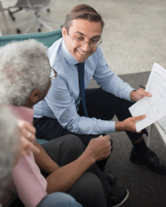 A professional man in a blue shirt and tie smiles, holding papers, as he discusses with two seniors in an office. The mood is friendly and collaborative.