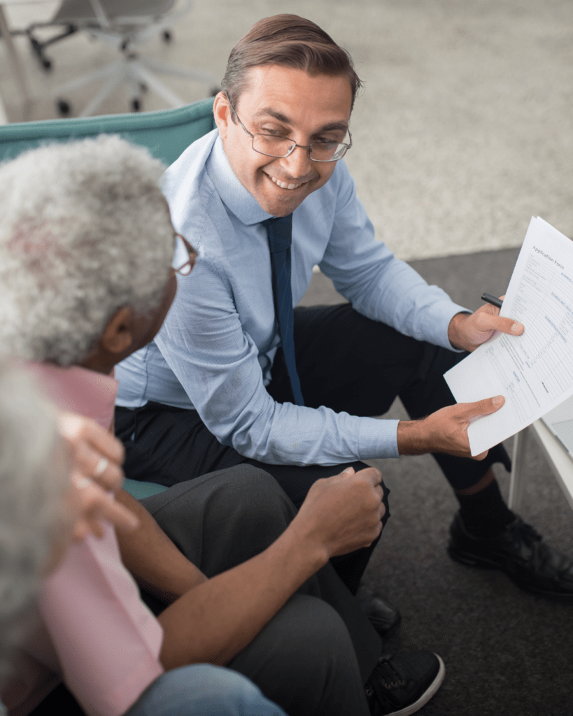 A professional man in a blue shirt and tie smiles, holding papers, as he discusses with two seniors in an office. The mood is friendly and collaborative.